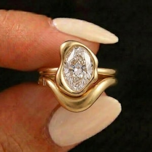May include: A gold ring featuring a central marquise-cut clear gemstone set within an organic, asymmetrical gold band. The metal has a smooth, polished finish that curves around the stone, creating a fluid, continuous shape on the finger.