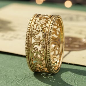 以下が含まれることがあります： 精巧なフィリグリーデザインのゴールドカラーのリング。リングは、渦巻く花のモチーフとビーズの縁取りの繊細なパターンが特徴です。リングは緑色の表面に置かれ、柔らかな照明とぼやけた背景があります。