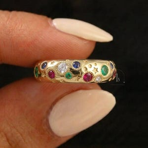 Op de afbeelding: Een gouden ring met een hemels ontwerp. De ring is versierd met verschillende gekleurde edelstenen, waaronder rood, blauw, groen en wit, geplaatst tussen ster- en stipmotieven. De ring wordt tussen twee vingers gehouden.