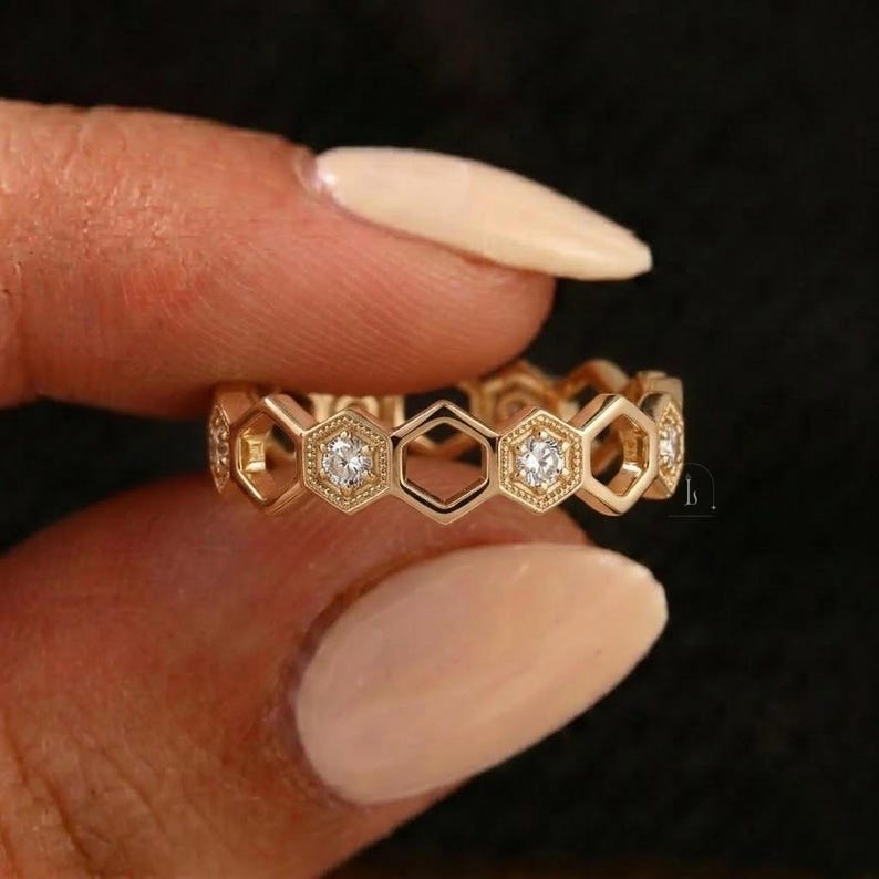 Pu&ograve; includere: Un anello color oro con un design a nido d'ape. L'anello presenta forme esagonali, alcune delle quali contengono piccole pietre preziose rotonde e trasparenti. L'anello &egrave; tenuto tra le dita su uno sfondo scuro.