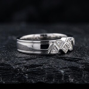 May include: A silver ring with a geometric design. The ring features a series of triangular and square-shaped diamond accents. The band has a polished finish with a grooved detail. The ring is displayed on a dark, textured surface.