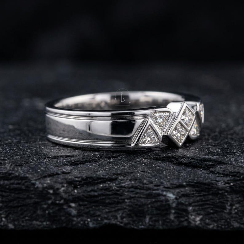 May include: A silver ring with a geometric design. The ring features a series of triangular and square-shaped diamond accents. The band has a polished finish with a grooved detail. The ring is displayed on a dark, textured surface.
