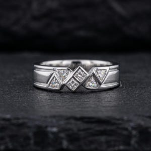 May include: A silver ring with a geometric design. The ring features square and triangular-shaped clear stones set in a band with a rectangular design. The ring is displayed on a dark surface.
