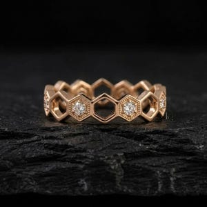 Pu&ograve; includere: Un anello in oro rosa con un design a nido d'ape. Ogni esagono presenta un piccolo diamante scintillante. L'anello &egrave; posto su uno sfondo scuro e strutturato, evidenziando i suoi dettagli intricati e il suo stile elegante.