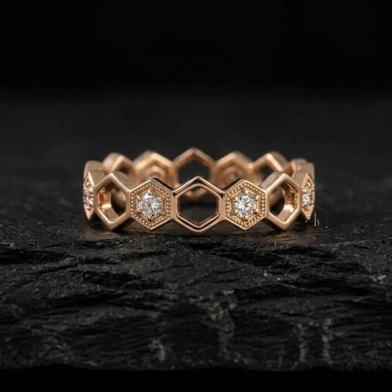 Pu&ograve; includere: Un anello in oro rosa con un design a nido d'ape. Ogni esagono presenta un piccolo diamante scintillante. L'anello &egrave; posto su uno sfondo scuro e strutturato, evidenziando i suoi dettagli intricati e il suo stile elegante.