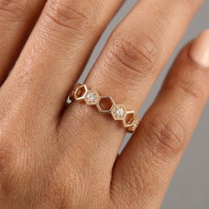 Pu&ograve; includere: Un anello color oro con un design a nido d'ape. L'anello presenta forme esagonali, ognuna contenente una piccola gemma scintillante. L'anello &egrave; indossato su un dito, mostrando i suoi dettagli intricati e il suo stile elegante.