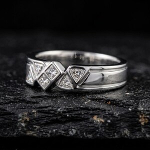 May include: A silver ring with a geometric design featuring square and triangular-cut gemstones. The ring has a polished finish and a band with a grooved detail. The gemstones are set in a repeating pattern across the top of the ring.