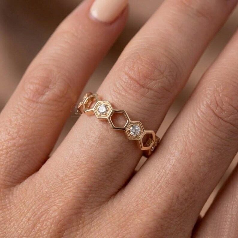 Pu&ograve; includere: Un anello color oro con un motivo a nido d'ape. Ogni sezione esagonale &egrave; bordata e incastonata con un piccolo diamante. L'anello &egrave; mostrato su un dito, evidenziando il suo design dettagliato e la sua maestria artigianale.