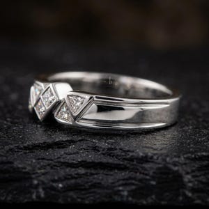 May include: A silver ring with a geometric design. The ring features a series of square and triangular-shaped clear stones set into the band. The band has a polished finish, reflecting light. The ring is displayed on a dark, textured surface.