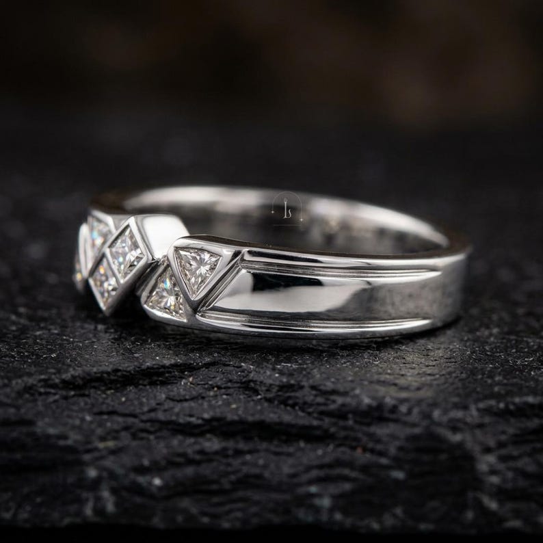 May include: A silver ring with a geometric design. The ring features a series of square and triangular-shaped clear stones set into the band. The band has a polished finish, reflecting light. The ring is displayed on a dark, textured surface.