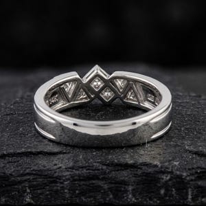May include: A silver ring with a geometric design featuring multiple square and triangular-shaped clear stones. The ring is set on a dark, textured surface, showcasing its polished metal and intricate details. The band is marked with "14K".