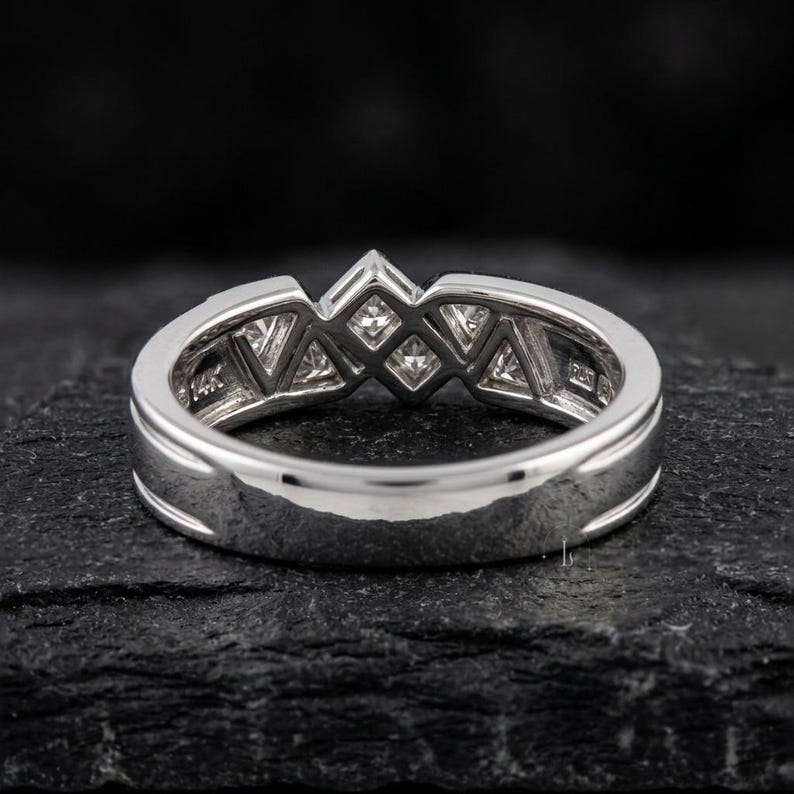 May include: A silver ring with a geometric design featuring multiple square and triangular-shaped clear stones. The ring is set on a dark, textured surface, showcasing its polished metal and intricate details. The band is marked with "14K".