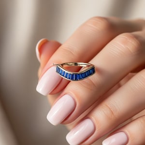 Peut inclure: Une bague en or avec un design incurvé, sertie de multiples pierres précieuses rectangulaires bleu foncé. La bague est présentée sur un fond doux et neutre, mettant en valeur les pierres bleues vibrantes.