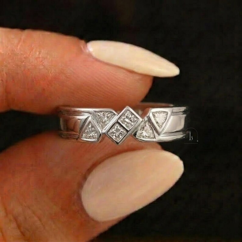 May include: A silver ring with a geometric design featuring square and triangular gemstones. The ring's band has a polished finish, and the gemstones are set in a repeating pattern. The ring is held by a person.