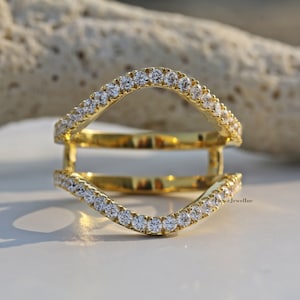 May include: A gold-colored ring guard with a double band design. The bands are lined with small, clear gemstones. The ring guard is designed to be worn with another ring. The background is a neutral color.