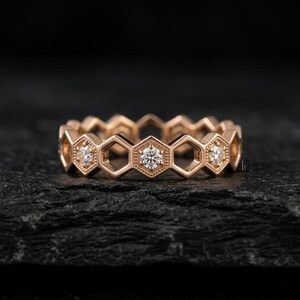 Pu&ograve; includere: Un anello in oro rosa con un design a nido d'ape. Ogni esagono presenta una piccola gemma rotonda e trasparente. L'anello &egrave; posto su uno sfondo scuro e strutturato, evidenziando i dettagli del gioiello.