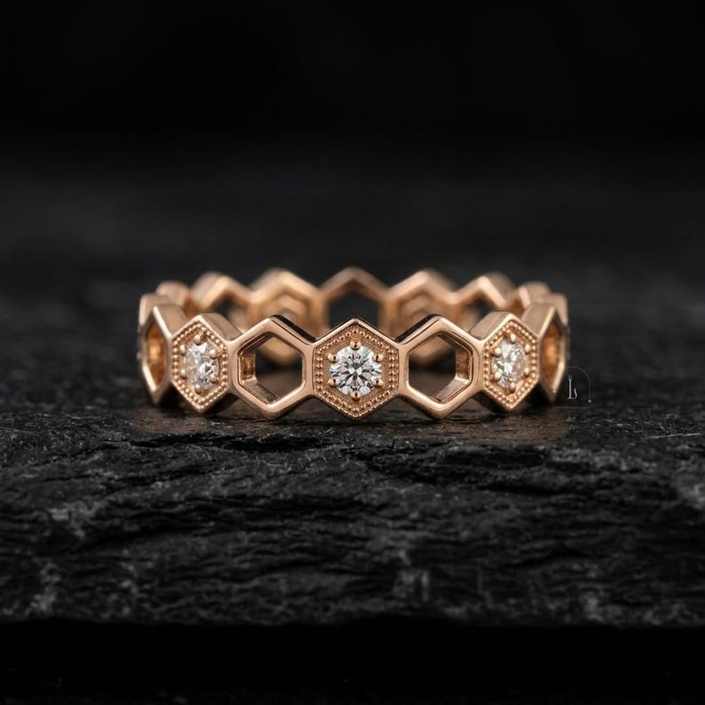 Pu&ograve; includere: Un anello in oro rosa con un design a nido d'ape. Ogni esagono presenta una piccola gemma rotonda e trasparente. L'anello &egrave; posto su uno sfondo scuro e strutturato, evidenziando i dettagli del gioiello.