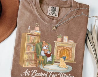 May include: A brown t-shirt with a snowman sitting in a chair by a fireplace reading a book. A dog sits next to the snowman. The text "All Booked For Winter" is printed on the shirt.