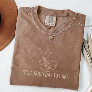 Peut inclure: Un t-shirt marron avec le texte "C'est un bon jour pour faire de la pâtisserie" et un graphique d'un bol à mélanger avec un fouet et des cœurs.