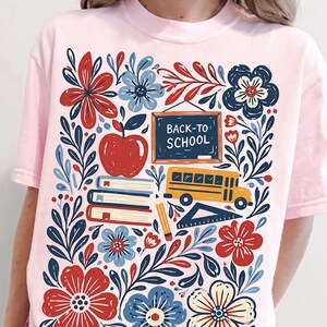 Peut inclure: Un t-shirt rose clair avec une impression graphique représentant une pomme rouge, un bus scolaire jaune, une pile de livres, un crayon, une règle et un tableau noir avec le texte BACK-TO-SCHOOL, entourés de motifs floraux rouges, bleus et crème.