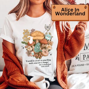 Peut inclure: T-shirt blanc avec un motif "Alice au pays des merveilles" repr&eacute;sentant des champignons, une chenille et d'autres personnages fantastiques. Le t-shirt comprend la citation, "It would be so nice if something made sense for a change!" et le texte "Alice in Wonderland Lewis Carroll".