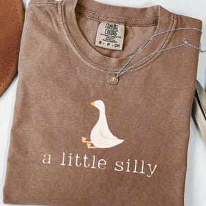 Peut inclure: Un t-shirt marron à manches courtes orné d'un motif représentant une oie blanche assise avec des pattes et un bec orange. Sous l'oie, le texte "a little silly" est inscrit en lettres blanches. Un collier en argent avec un pendentif repose dessus.