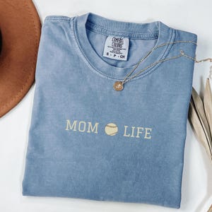 Peut inclure: Un t-shirt bleu clair avec le texte "MOM LIFE" en blanc et un graphique de baseball au milieu. Le t-shirt a un aspect vieilli et est fait d'un matériau doux et confortable.