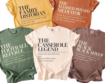 Puede incluir: Cinco camisetas de manga corta y cuello redondo en amarillo, naranja, verde, crema y marrón. Cada camiseta presenta texto negro en estilo de definición de diccionario con títulos como The Family Historian, The Mashed Potato Mediator, The Football Referee, The Casserole Legend y The Black Black Sheep. El texto inferior dice 16 DESIGNS TO CHOOSE FROM.