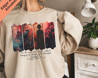 May include: Beige sweatshirt featuring a graphic design with imagery from Edgar Allan Poe's poem, "Annabel Lee." The design includes a raven, a silhouette of a person, and floral elements. The text "For the man never became, without bringing me dreams of the beautiful" is also included.