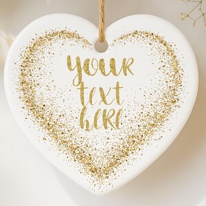 May include: A white heart-shaped ornament with gold glitter accents and the text "your text here" in gold script. The ornament has a hole at the top for hanging with a natural twine string. The design is simple and elegant.