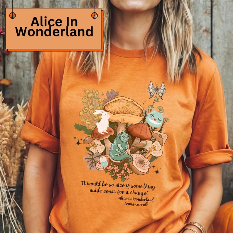 Peut inclure: T-shirt orange avec un motif "Alice au pays des merveilles". Le graphisme comprend des champignons, un lapin blanc, une chenille et un chat du Cheshire. La citation "It would be so nice if something made sense for a change" est incluse.