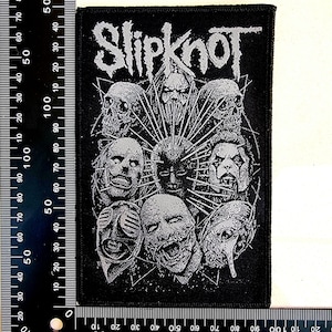 Può includere: Toppa rettangolare nera con il nome della band "Slipknot" in un font gotico. La toppa presenta una grafica in scala di grigi di più volti stilizzati con una figura centrale che irradia punte, evocando un'estetica dark heavy metal.