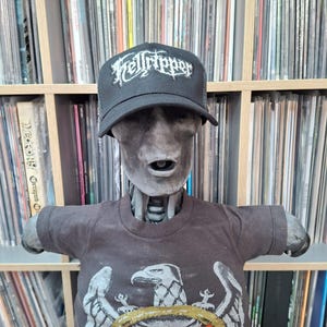 May include: A black trucker hat with the text "Helltripper" in white lettering is worn by a gray mannequin. The mannequin is also wearing a dark grey t-shirt with an eagle graphic. The background shows a shelf filled with vinyl records.