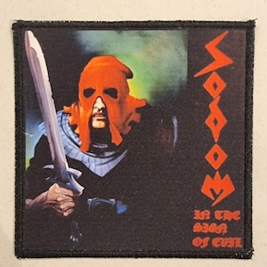 May include: Square patch featuring a figure in an orange hood holding a sword. The patch has the word "SODOM" in red and the text "IN THE SIGN OF EVIL". The background is dark with a black border.