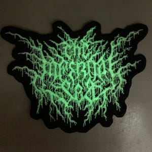 May include: A black and green embroidered patch with the text "The Black Dahlia Murder" in a gothic font. The patch glows in the dark.
