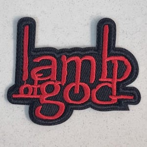Könnte beinhalten: Gestickter Aufnäher mit dem Schriftzug "Lamb of God" in Rot auf schwarzem Grund. Der Text ist in einer stilisierten Schriftart gehalten. Der Aufnäher hat eine strukturierte Optik.