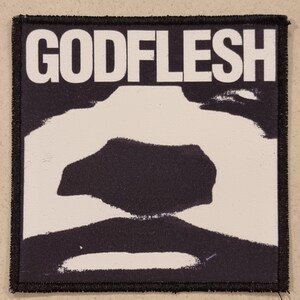 May include: A square patch with the word "GODFLESH" in bold white letters at the top. The patch has a black border and a black and white abstract design in the center.