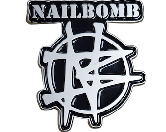 May include: A black and silver metal pin with the word "NAILBOMB" in white block letters at the top. The pin features a stylized, geometric design in black and silver.