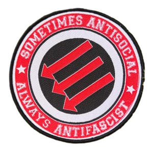 Op de afbeelding: Ronde patch met een zwart midden, witte rand en rode buitenring. De tekst "SOMETIMES ANTISOCIAL" en "ALWAYS ANTIFASCIST" zijn rood. Drie rode pijlen wijzen naar beneden in het midden.