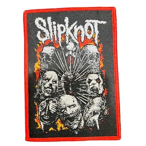 May include: Black rectangular patch with a red border. The patch features the band name "Slipknot" in white, above a graphic of several grotesque faces and skulls. The background is black with orange and yellow flames.