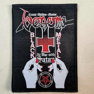 Può includere: Un cerotto bianco e nero con il nome della band "Venom" in un font stilizzato. Il testo "Black Metal" è su entrambi i lati di una croce rossa. Il testo "At War with Satan" è sotto la croce. Il cerotto presenta due mani che tengono la croce e un pentagramma con una testa di capra al centro.