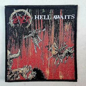 Könnte beinhalten: Ein schwarz-weißes Patch mit dem Bandnamen "Slayer" und den Worten "Hell Awaits" in Rot. Das Patch zeigt einen stilisierten Schädel mit Hörnern vor einem Hintergrund aus Flammen.