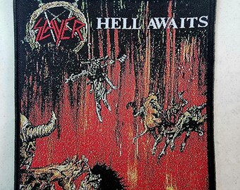 May include: A black and white patch featuring the band name "Slayer" and the words "Hell Awaits" in red. The patch depicts a stylised skull with horns against a backdrop of flames.
