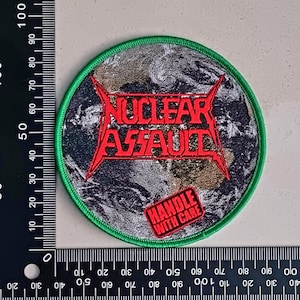 Könnte beinhalten: Ein runder grüner Patch mit einem Schwarzweißbild der Erde. Der Text "NUCLEAR ASSAULT" ist rot und der Text "HANDLE WITH CARE" ist rot und weiß.