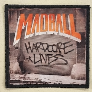 May include: Embroidered patch with the word "MADBALL" in orange block letters above the words "HARDCORE LIVES" in black graffiti-style lettering. The background shows a person walking in front of a metal door.