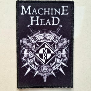 Könnte beinhalten: Schwarzer rechteckiger Aufnäher mit dem weißen Text "MACHINE HEAD" über einem detaillierten Emblem. Das Emblem zeigt gekreuzte Schwerter, Löwen und ein zentrales geometrisches Design. Der Aufnäher hat einen schwarzen Rand.