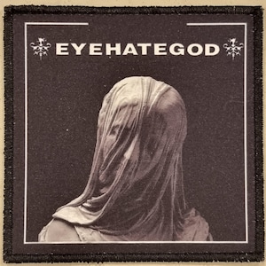 May include: A black and white patch with the band name "EYEHATEGOD" in white capital letters. The patch features a sepia-toned image of a veiled figure. It has a black border and a white inner frame.