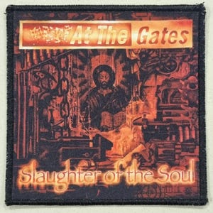 Puede incluir: Parche cuadrado con borde negro con el texto "At The Gates" encima de "Slaughter of the Soul". La imagen muestra una figura religiosa y maquinaria industrial en tonos naranja y marrón.