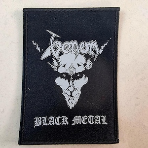 Op de afbeelding: Zwart-witte patch met de bandnaam "Venom" in een gestileerd lettertype. De patch toont een demonische figuur met hoorns en een pentagram op zijn voorhoofd. De tekst "Black Metal" is onder de figuur gedrukt.