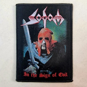 May include: Black rectangular patch with the band name "Sodom" in white and red. The image shows a figure in a red mask holding a sword, with the text "In the Sign of Evil" beneath. The patch is for clothing.